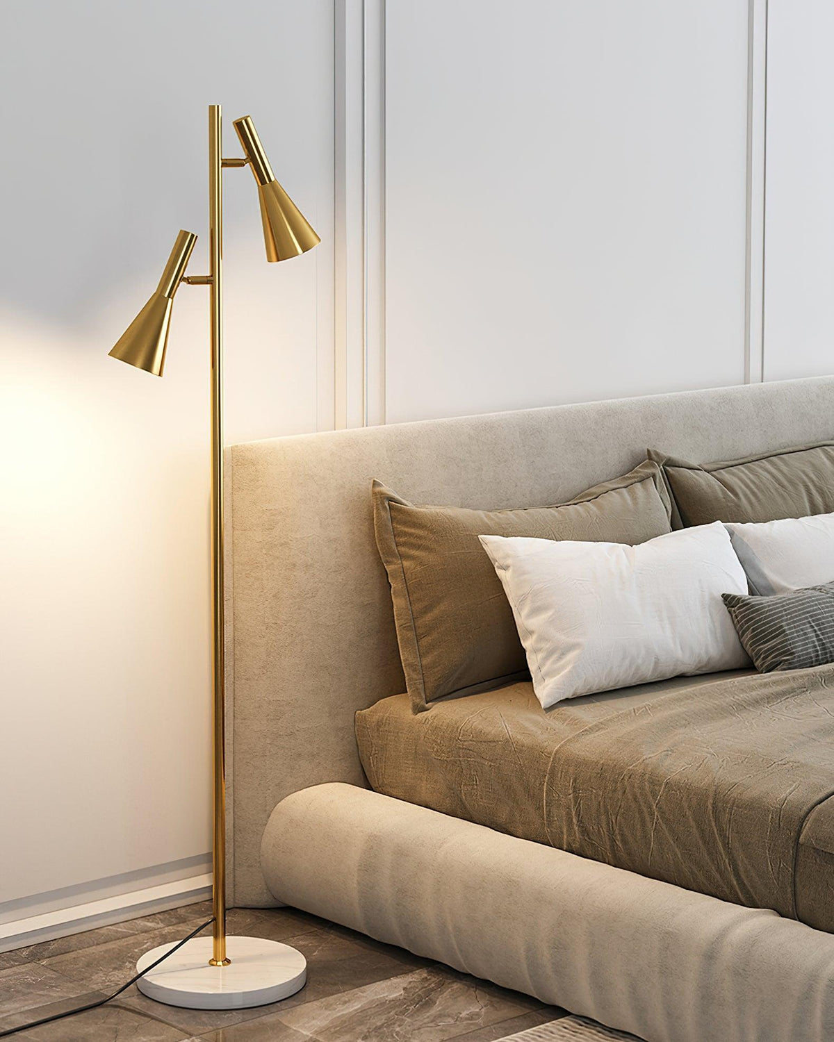 A beige upholstered bed with white pillows sits beside a brass Clérachic floor lamp glowing warm on its marble base.