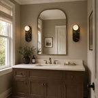 Two dark Clérachic sconces flank an arched mirror near a window, with a small candle set on the marble counter.