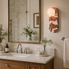 The Clérachic sconce glows beside a gilt mirror and sink, with a soap dispenser and dried flowers on the counter.