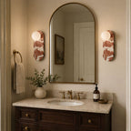 Two Clérachic sconces flank an arched mirror above a dark wood vanity, their marble bodies warmly lit in a bathroom.