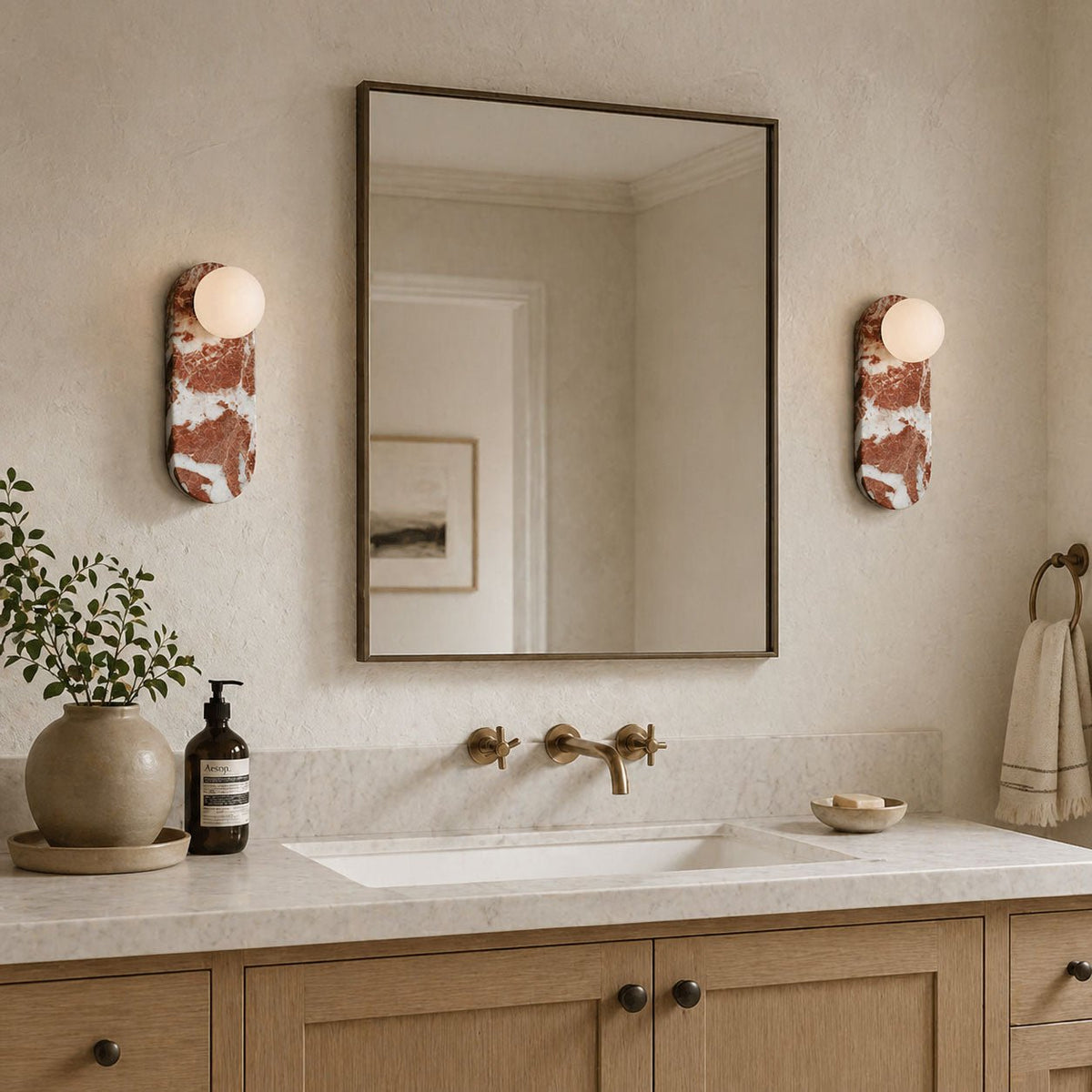 In a light oak bathroom, two Clérachic sconces frame a square mirror above a marble countertop and brass taps.