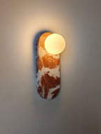 An angled close-up of the lit Clérachic sconce in red marble, casting a soft shadow across the wall behind it.