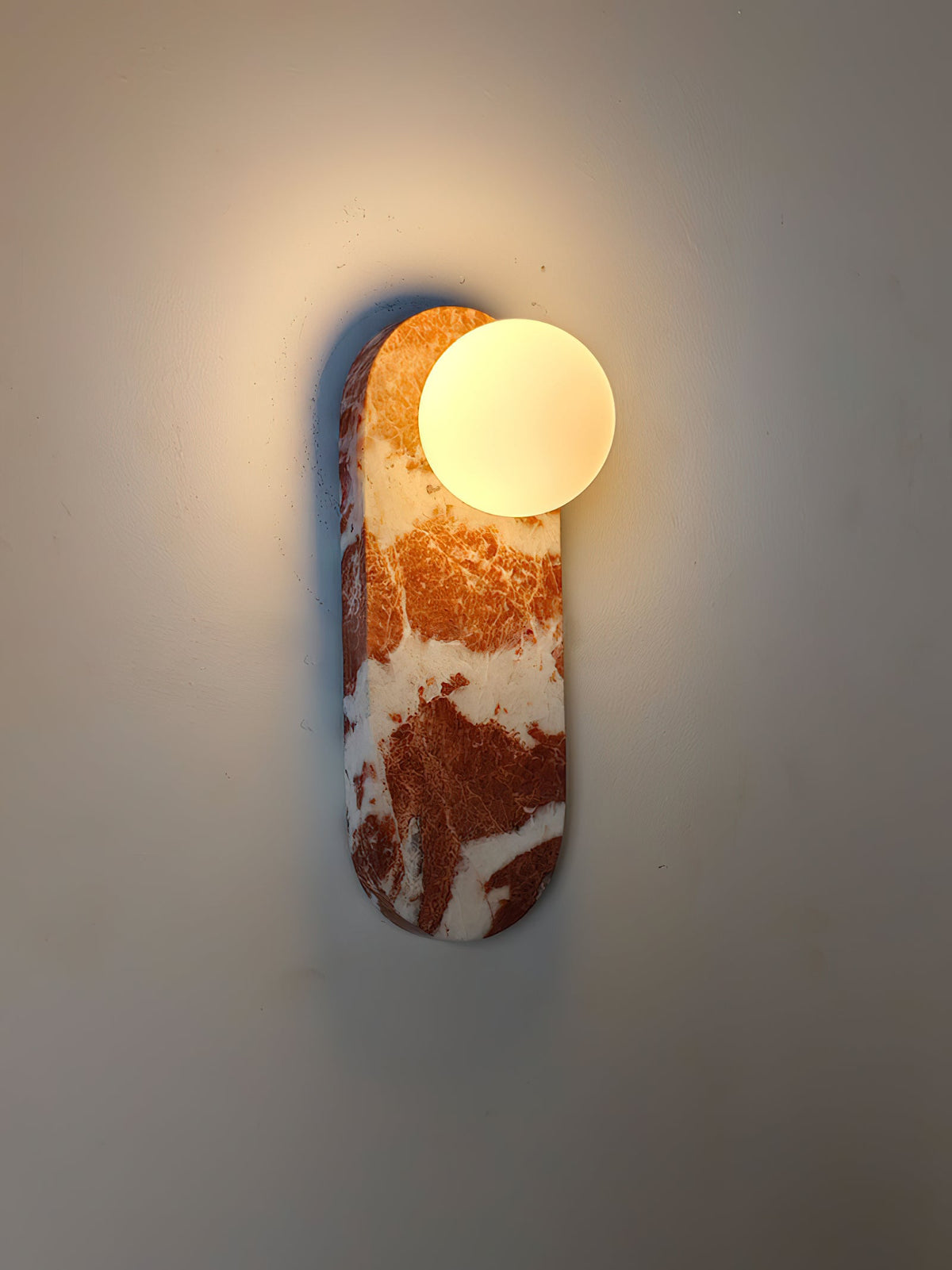 An angled close-up of the lit Clérachic sconce in red marble, casting a soft shadow across the wall behind it.