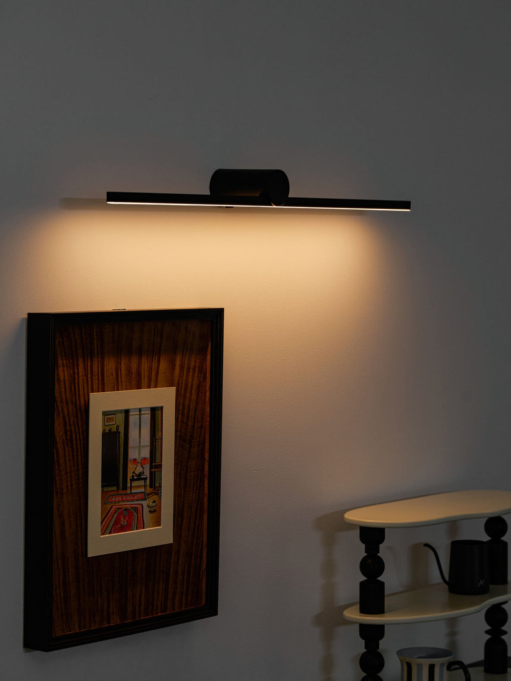 Angled view of a lit Clérachic wall light throwing warm light down a wood-framed picture.