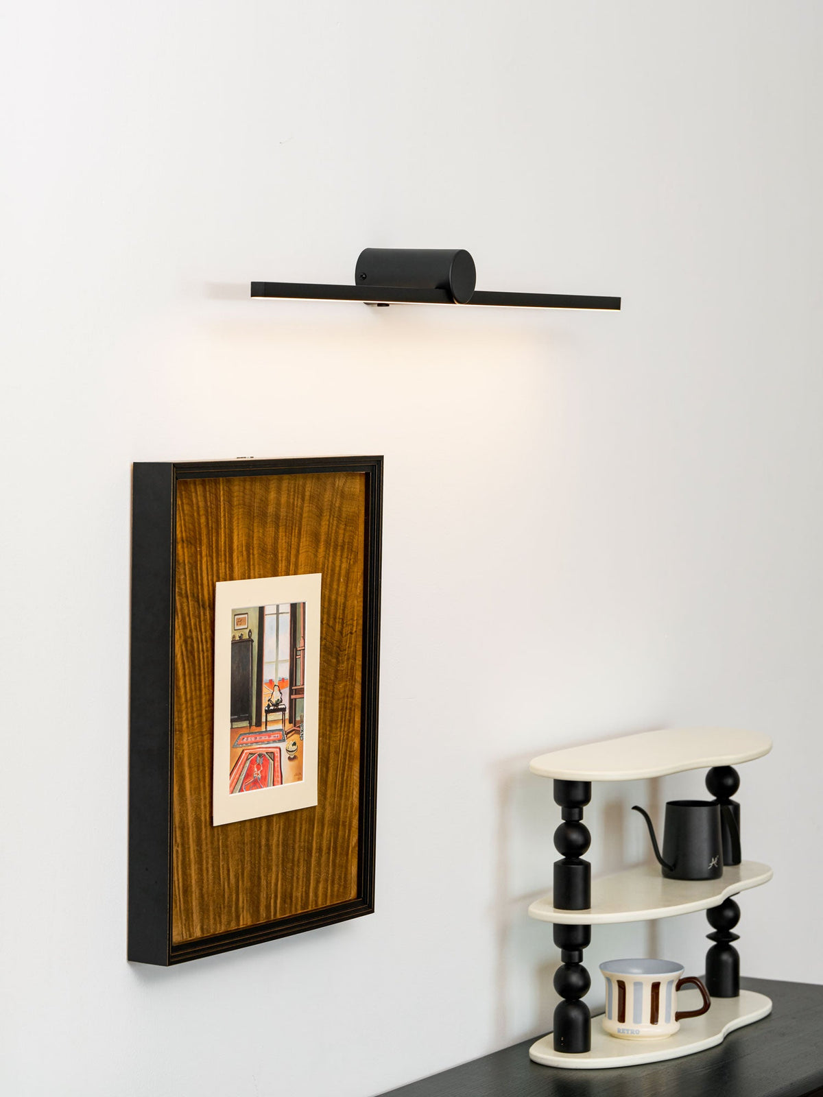 A lit Clérachic wall light glows above a black-framed print and small ladder shelf.