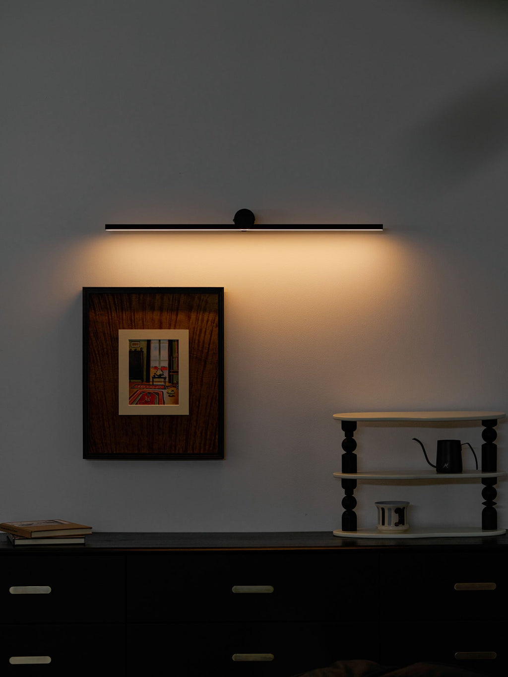 A lit Clérachic wall light glows above a black dresser, framed still-life print, and small ladder shelf.