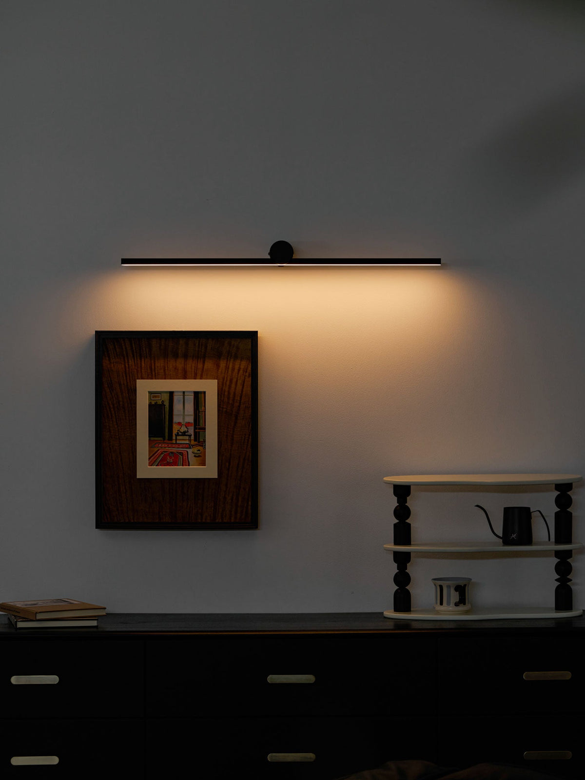 A lit Clérachic wall light glows above a black dresser, framed still-life print, and small ladder shelf.