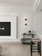 Two Clérachic alabaster wall lights stacked on a panelled white wall beside a media unit, near a bar table and stools.