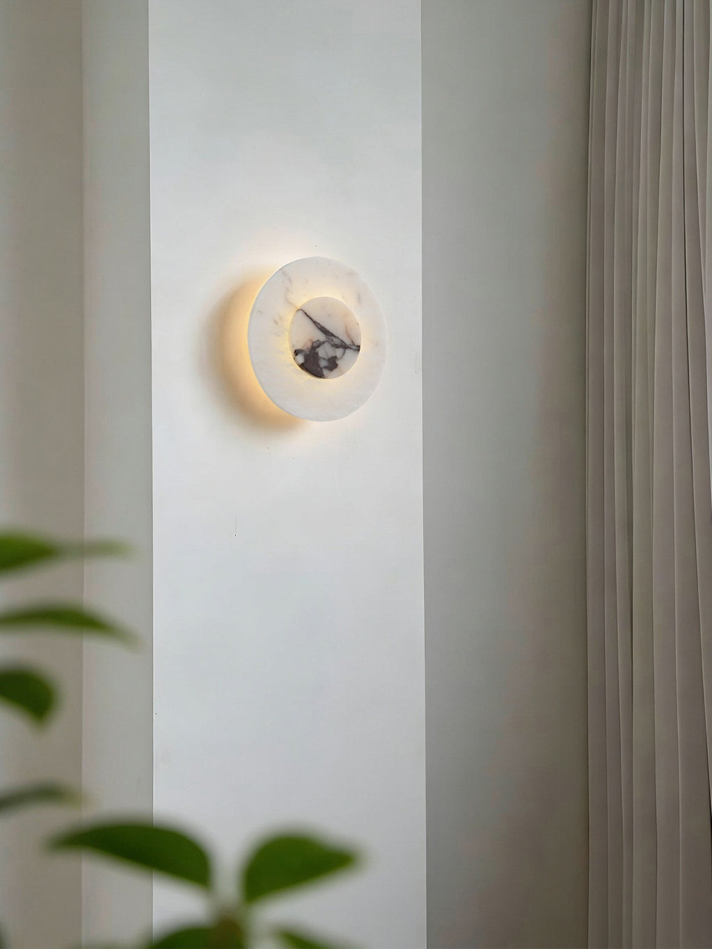 Another close-up of the lit Clérachic alabaster disc wall light on a wall pillar, framed by curtain fabric and green leaves.