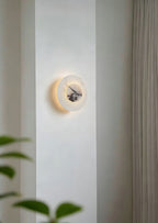 Close-up of a lit Clérachic alabaster disc wall light on a wall pillar, its dark marbled centre glowing against blurred leaves.