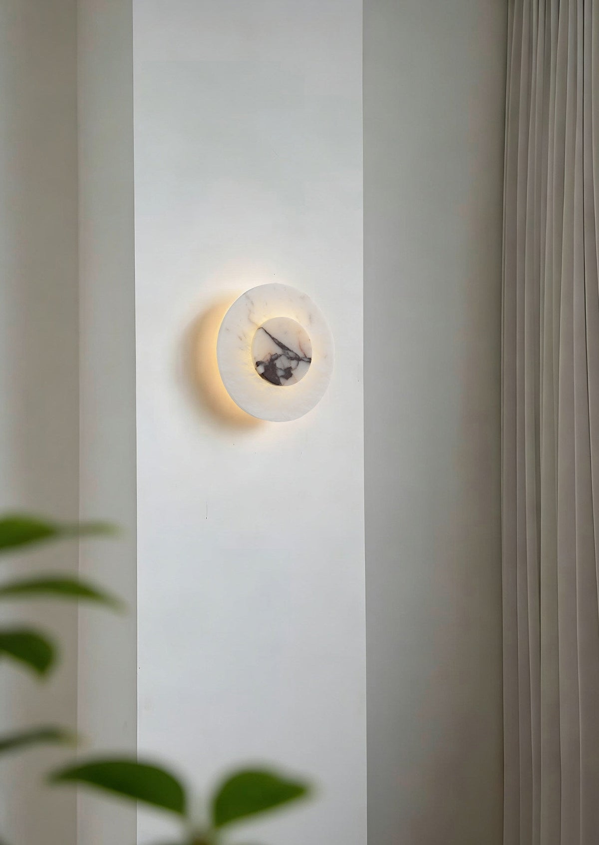 Close-up of a lit Clérachic alabaster disc wall light on a wall pillar, its dark marbled centre glowing against blurred leaves.
