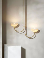 Clérachic two-arm Lunaris pendant lit above a white plinth, warm light pooling from both alabaster bowls onto the plaster wall.