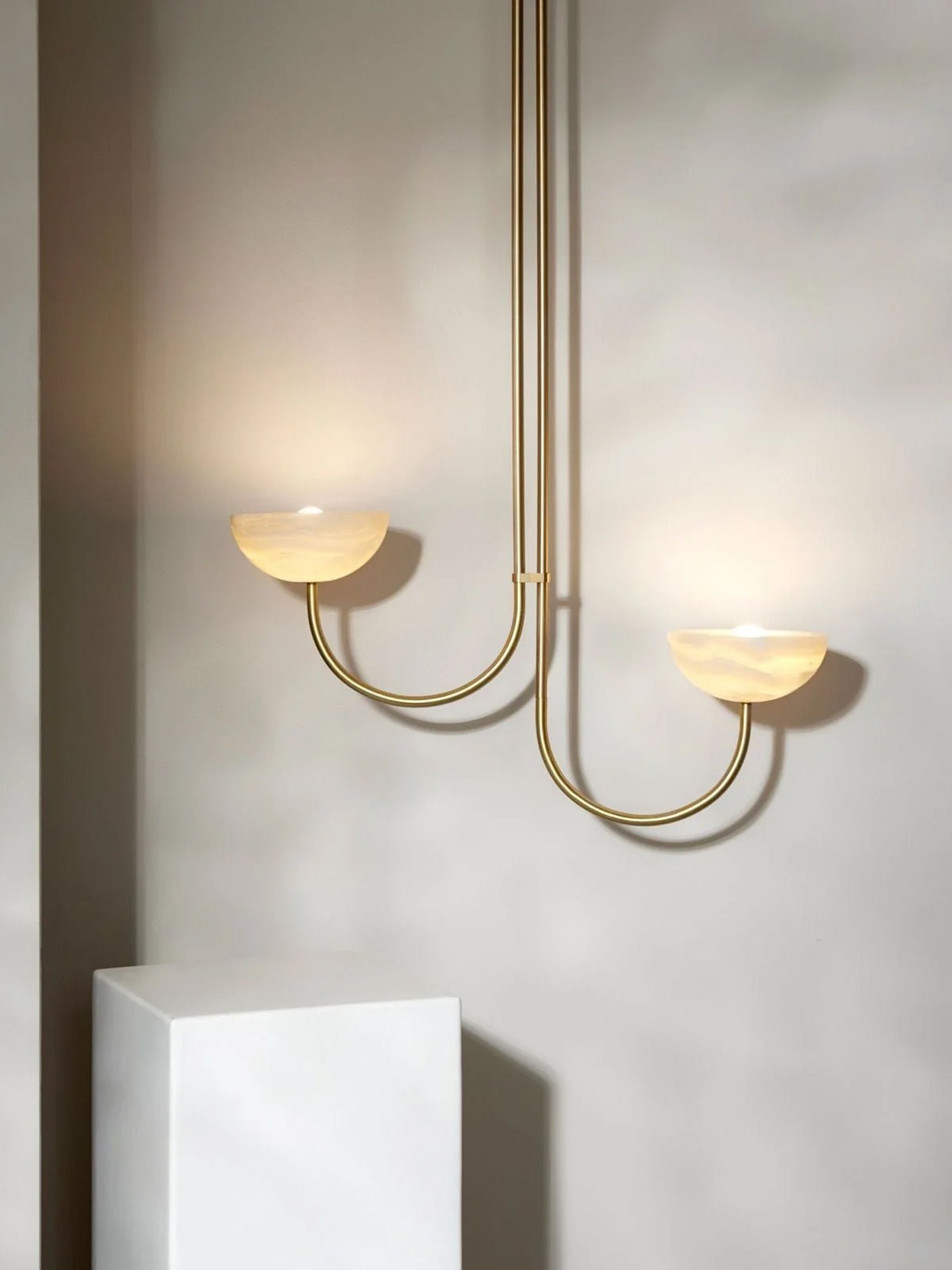 Clérachic two-arm Lunaris pendant lit above a white plinth, warm light pooling from both alabaster bowls onto the plaster wall.