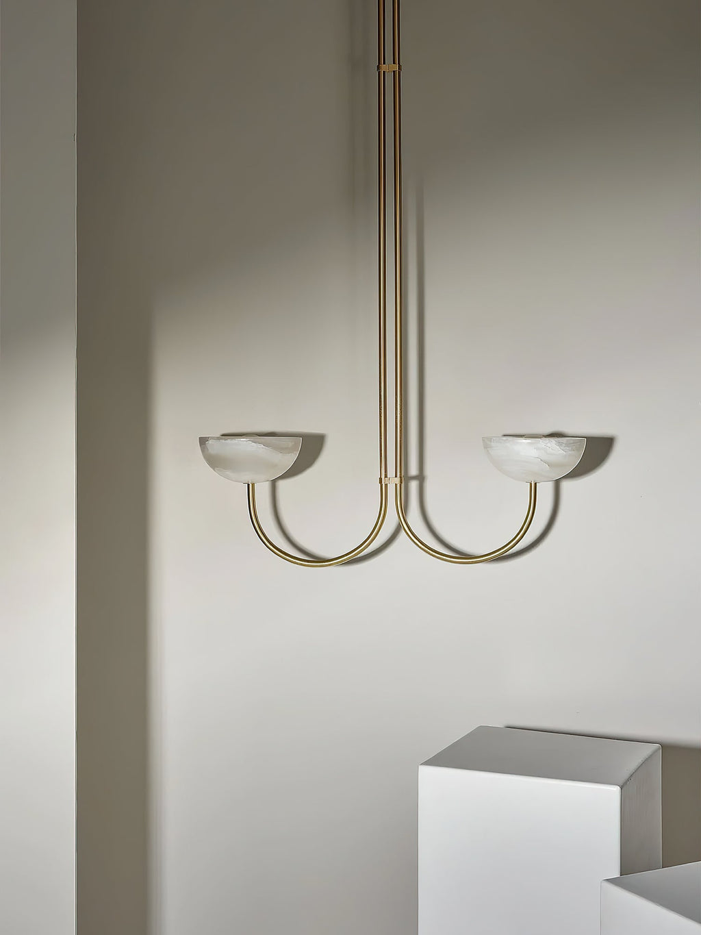 Clérachic brass Lunaris pendant with white alabaster bowls hanging in a plaster-walled room beside a white plinth.