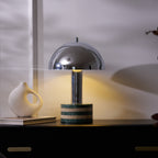 Wider view of the Clérachic table lamp lit on the dark cabinet, the ceramic vase and stacked books beside its marble base.