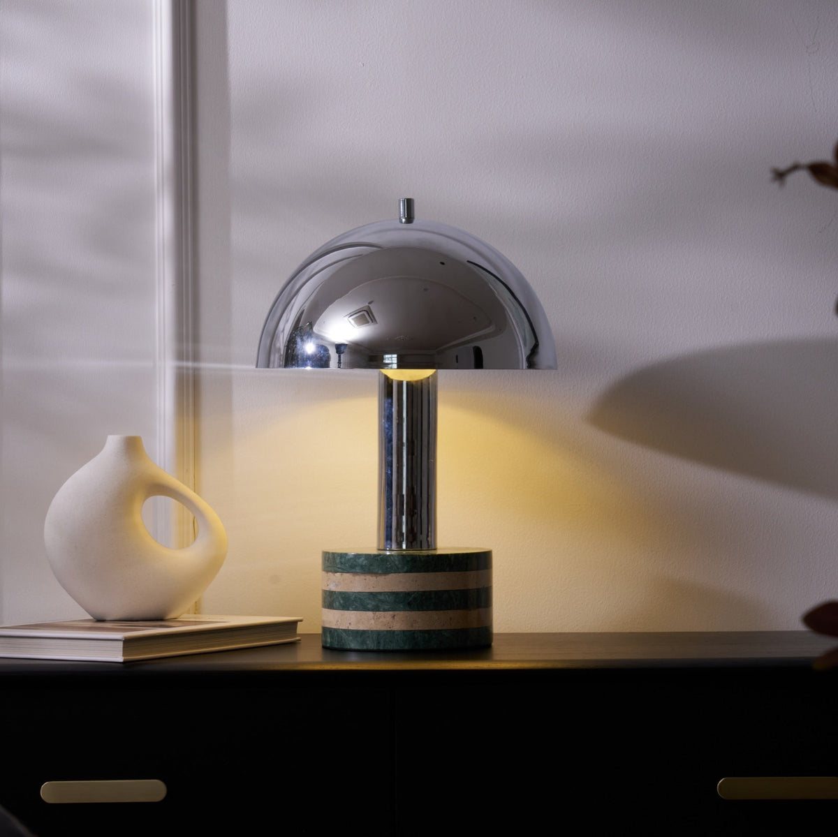 Wider view of the Clérachic table lamp lit on the dark cabinet, the ceramic vase and stacked books beside its marble base.