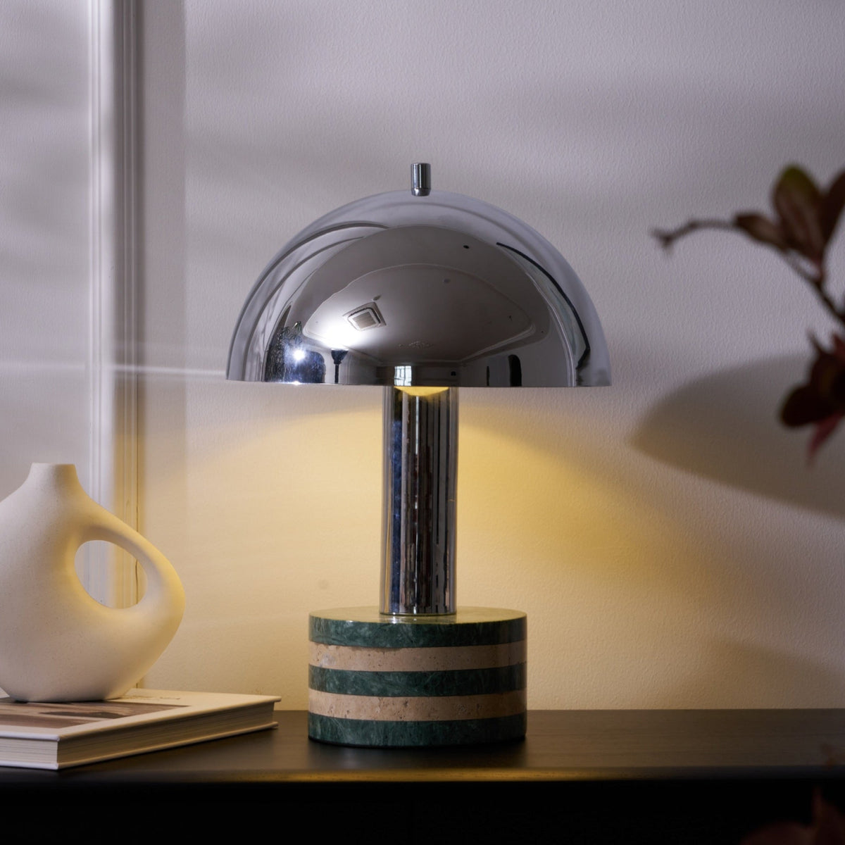 Medium crop of the lit Clérachic lamp with the vase and books to its left, warm light spilling across the cabinet top.