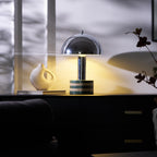 Wide shot of the lit Clérachic lamp on the cabinet, curtain shadows crossing the wall above the vase and books.