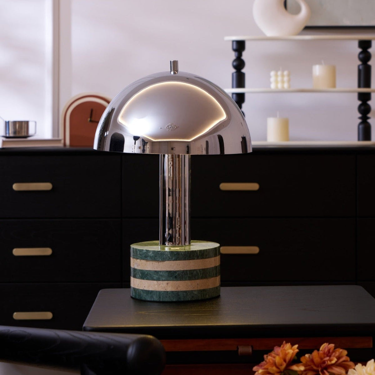 Clérachic lamp lit on a dark dresser, its dome rim glowing beside a turned-wood shelf holding candles and a clock.