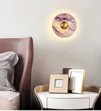 A lit Clérachic purple marble wall light hangs above a brown leather headboard, beside a round nightstand and framed photos.