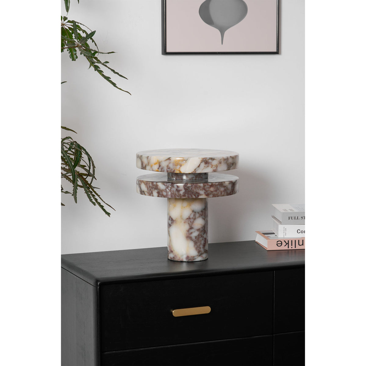 Clérachic lamp unlit beneath a pink abstract print, a branch reaching into frame beside the stacked marble discs.