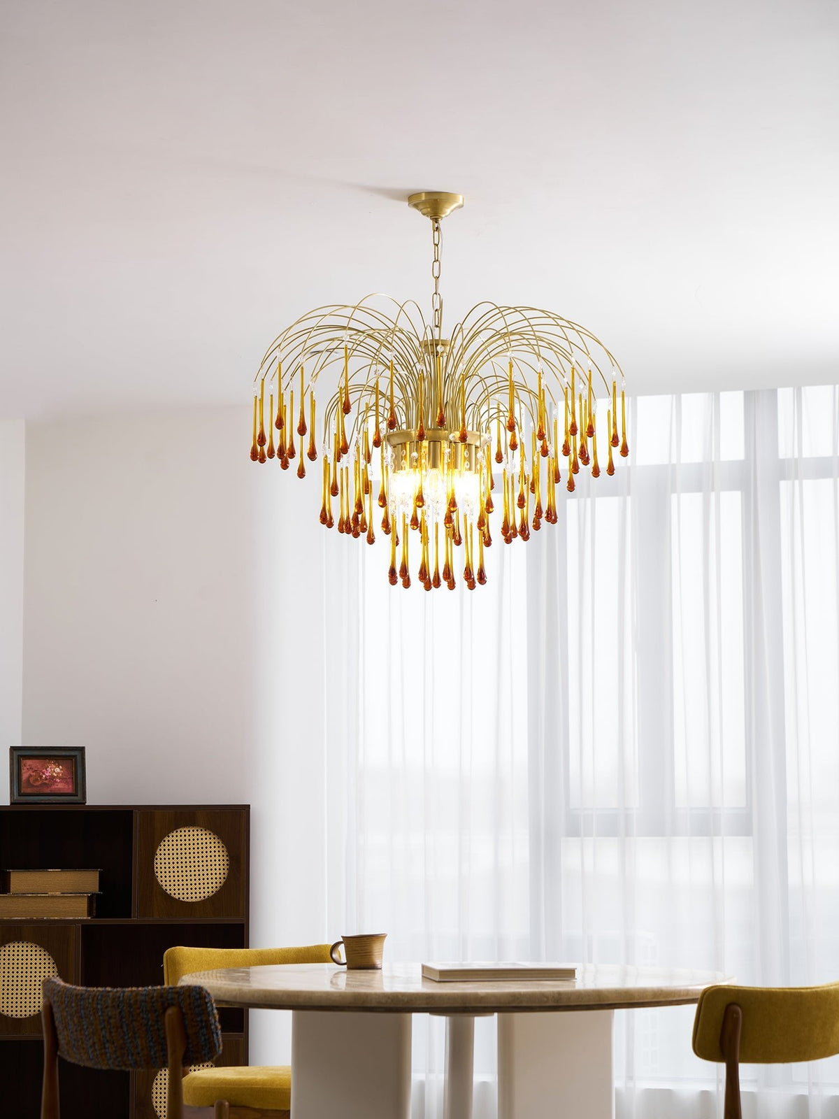 This Clérachic pendant's caramel crystal drops hang above a mustard-yellow chair, sheer curtains lit brightly behind it.