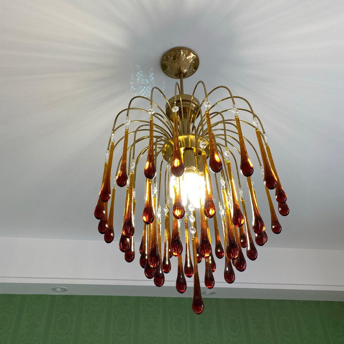 Dramatic sunburst rays cross the ceiling above this Clérachic chandelier, lit warm over a green-toned floor.