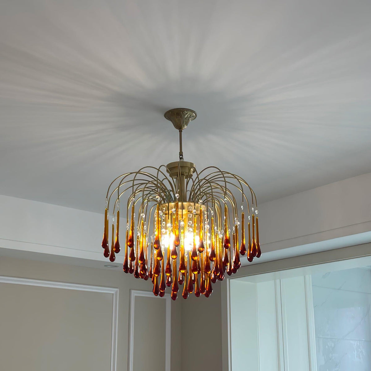 Dramatic light rays radiate across the ceiling above this Clérachic pendant, its caramel crystal drops glowing warm.