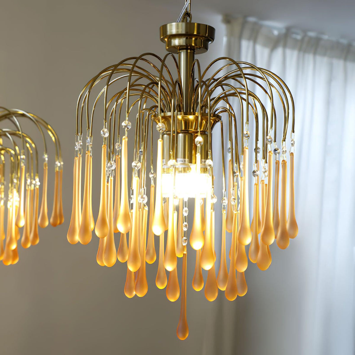 Looking up into this Clérachic chandelier, its lit bulbs sit among caramel and clear crystal drops beside a sheer curtain.