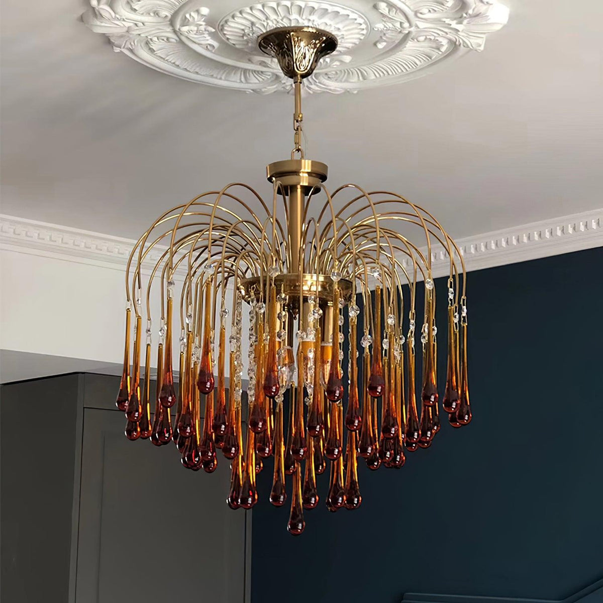 An ornate ceiling medallion sits above this Clérachic chandelier, its caramel and clear crystal drops set against a dark teal wall.