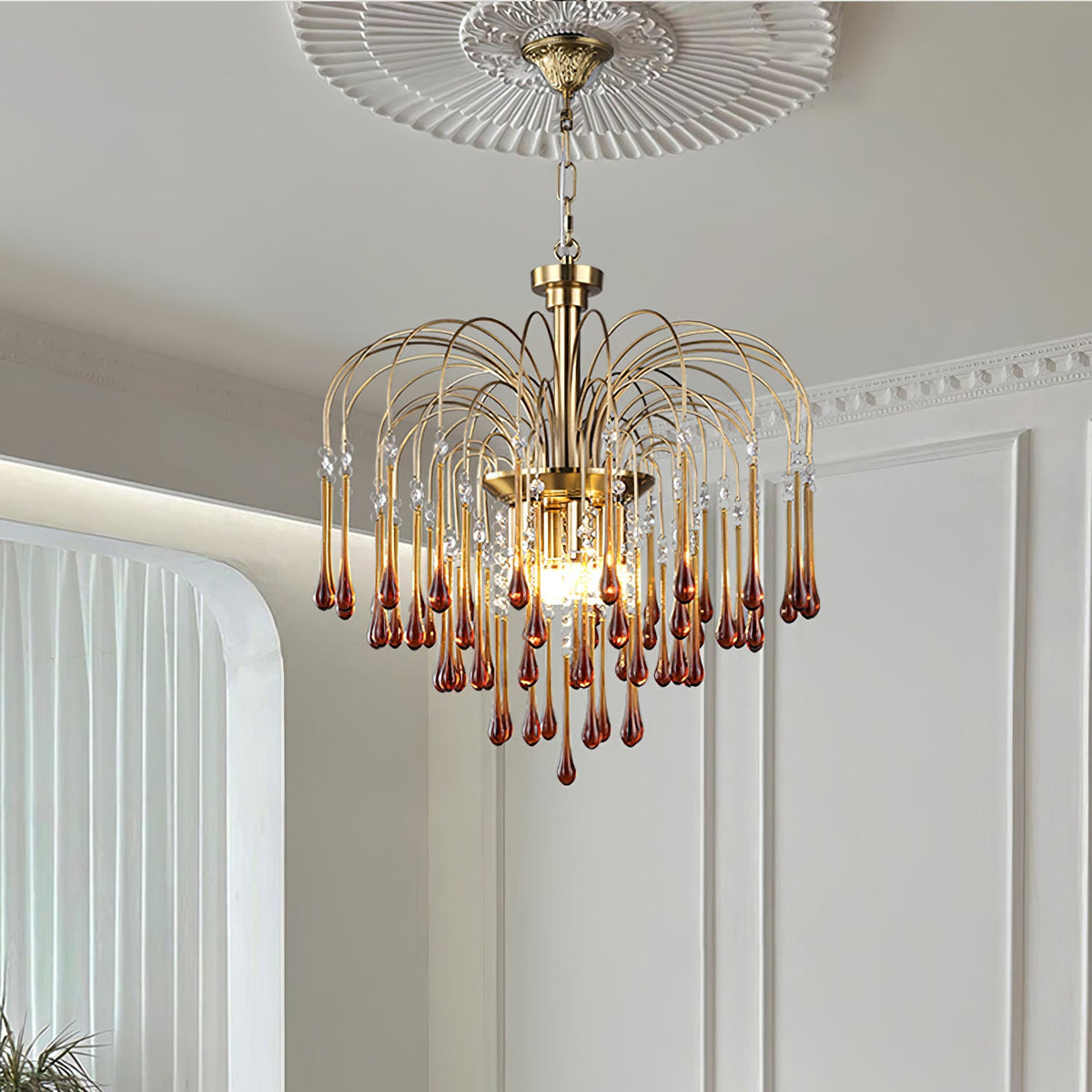 An ornate ceiling rose sits above this Clérachic pendant, its caramel and clear crystal drops lit beside a sheer curtain.
