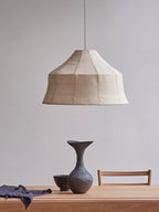 A faceted woven Clérachic pendant sits on a plain background, styled above a dark ceramic vase and blue cloth.