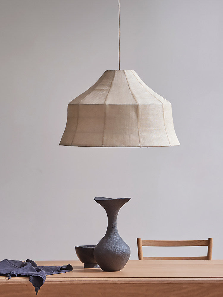 A faceted woven Clérachic pendant sits on a plain background, styled above a dark ceramic vase and blue cloth.