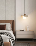 A travertine sail Clérachic pendant glows beside a tan leather headboard, a nightstand holding books and a small sculpture.