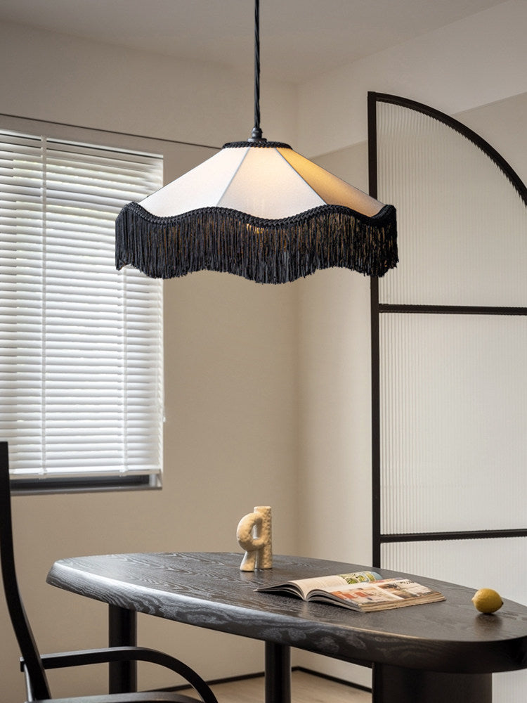 Above a dark wood table beside an arched black metal divider, this Clérachic pendant glows warm with its fringed hem.