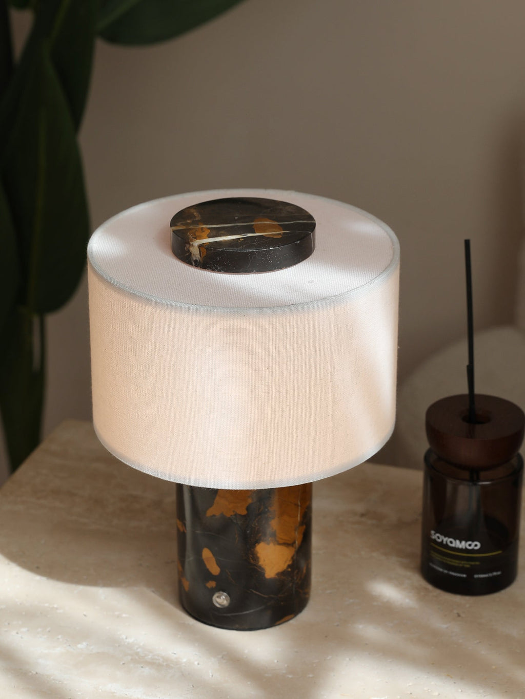 The Clérachic table lamp's cream shade and black-and-gold base sit beside a diffuser bottle on a sunlit surface.