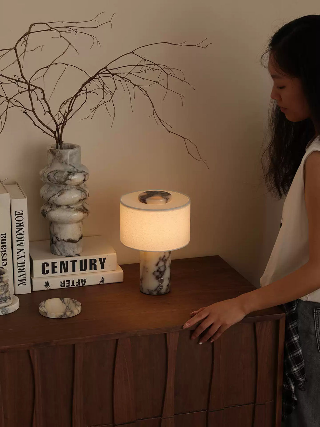 A woman glances down at the Clérachic table lamp glowing on a wooden sideboard beside the marble vase and books.