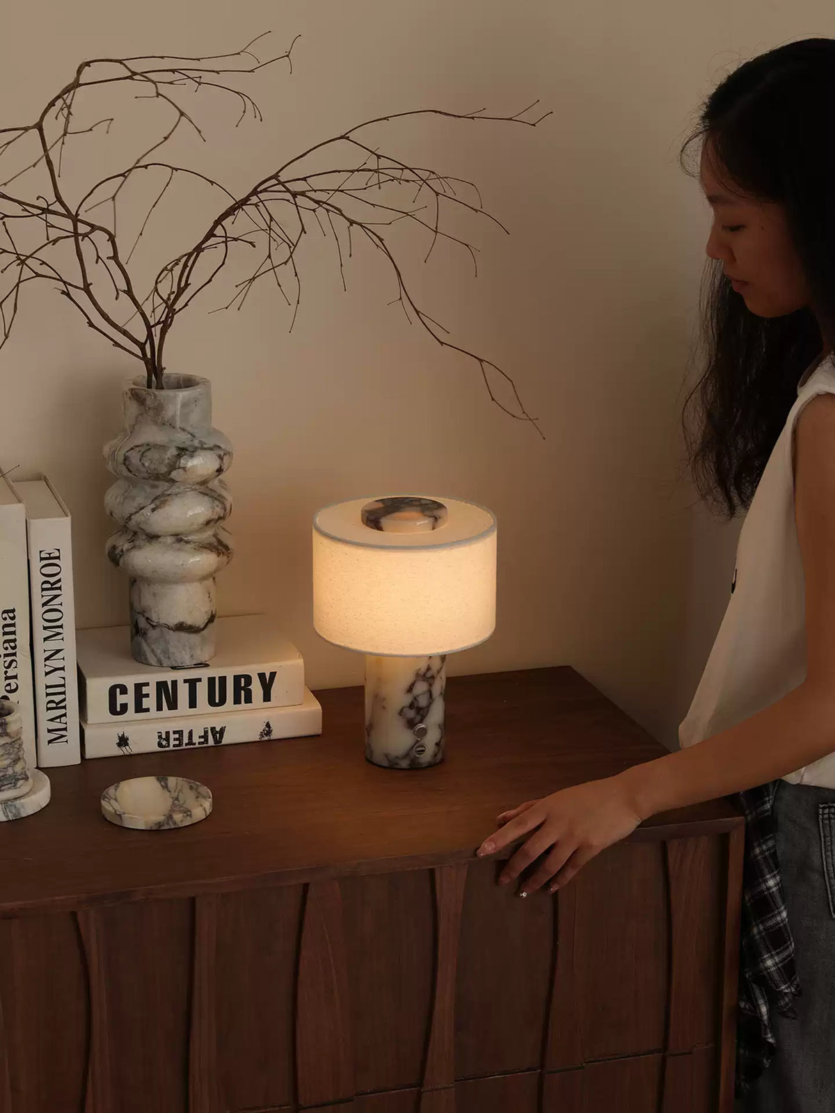 A woman glances down at the Clérachic table lamp glowing on a wooden sideboard beside the marble vase and books.