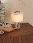 A hand reaches toward the Clérachic table lamp's base on a wooden sideboard, with a marble vase and books close by.