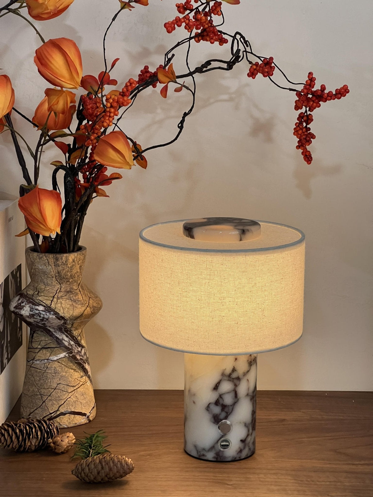 The Clérachic table lamp glows on a wooden table beside a rustic vase of orange berry branches and a pinecone.