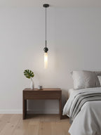 A single black Clérachic pendant light hangs above a wood nightstand holding a small vase and cup beside a bed with white linen.