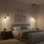 Three black Clérachic pendant lights at staggered heights glow above a bed with a tufted grey headboard and framed wall art.