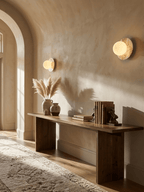 Two lit Clérachic marble disc sconces flank a plastered archway hallway, above a wood bench with vases and stacked books.