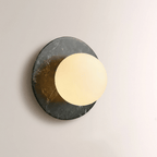 Close-up of a Clérachic wall sconce in dark marble, its opal globe glowing warm gold against the stone's mottled surface.