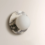 Grey-veined white marble surrounds this Clérachic wall sconce, the frosted glass globe unlit against the natural stone pattern.