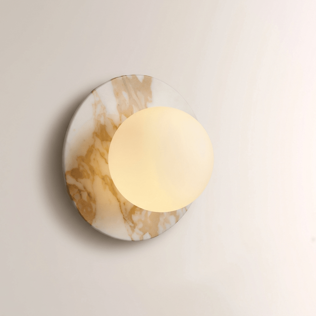 Close-up of the gold-veined marble Clérachic wall sconce with its opal globe lit warm, casting a soft glow across the disc.
