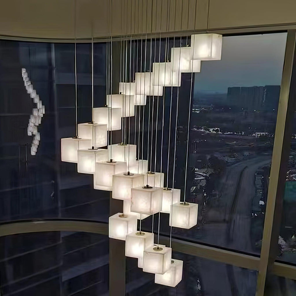 Alabaster cube pendants cascading in a spiral down a stairwell beside a floor-to-ceiling window at dusk.