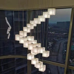 Alabaster cube pendants cascading in a spiral down a stairwell beside a floor-to-ceiling window at dusk.