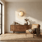 A lit Clérachic marble disc sconce hangs above a walnut sideboard and tan leather armchair in a sunlit, minimalist room.