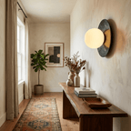 In a narrow hallway, a lit Clérachic marble disc sconce hangs above a console table with a potted fig tree and a patterned rug.